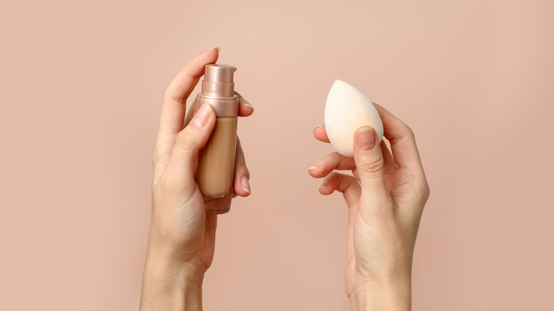 hand holding a foundation bottle and the other hand holding a makeup sponge